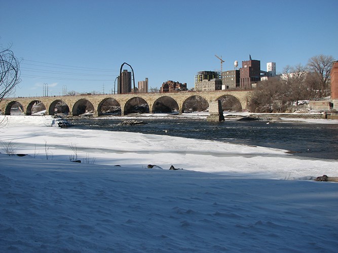 A stone-arch bridge over a partially frozen river against an urban skyline.