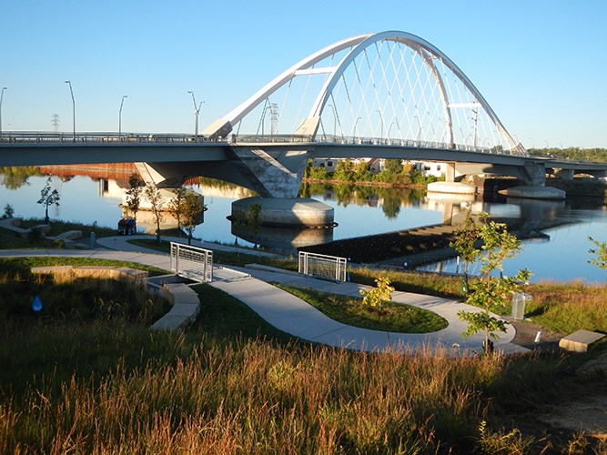 A modern arched bridge spans a calm river above a landscaped park path with grass, trees, and clear blue sky.