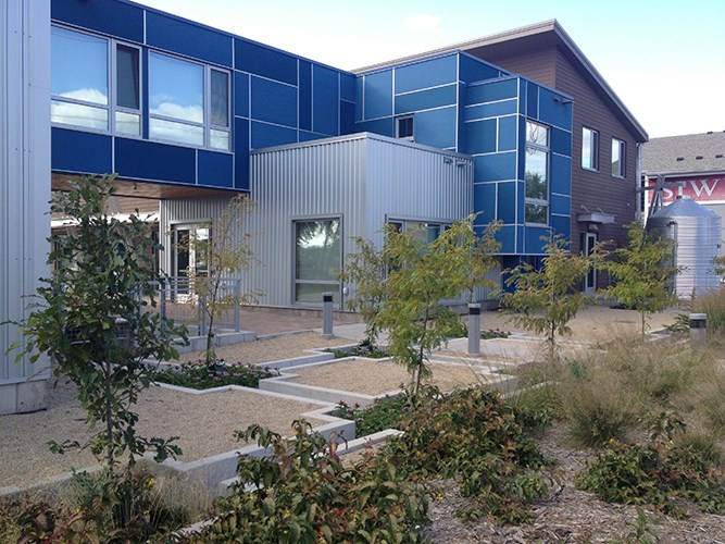A modern building with blue and silver geometric panels is surrounded by a landscaped courtyard featuring gravel paths, young trees, and low plantings.