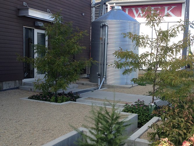 A metal rainwater tank stands beside a modern building amid a gravel courtyard with young trees and low planter beds.