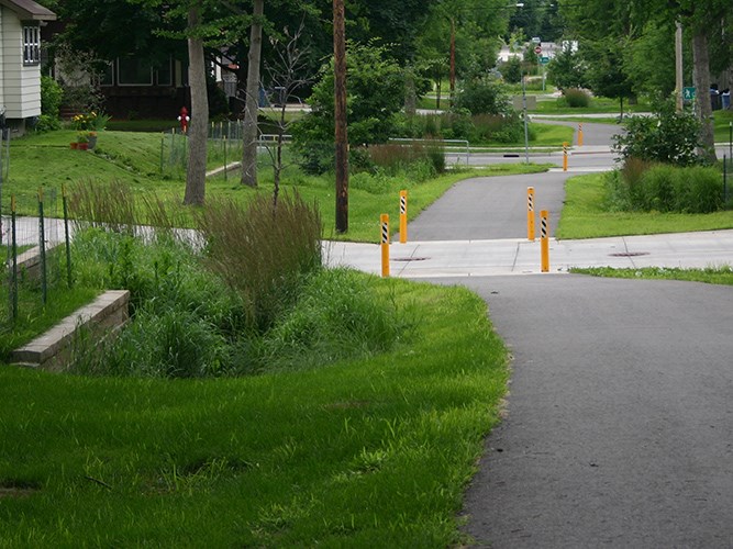 A paved trail curves through a grassy neighborhood area and crosses a sidewalk marked by several bright yellow bollards.