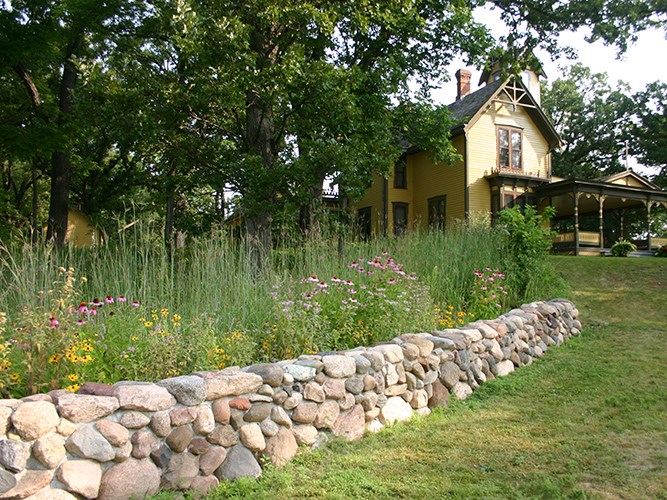 A stone retaining wall borders a garden of tall grasses and wildflowers in front of a yellow historic-style house shaded by large trees.