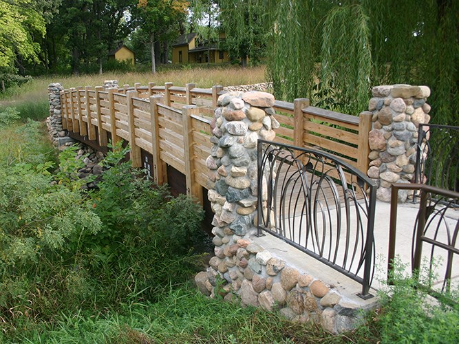 A wooden bridge with stone pillars and metal railings spans a lush, overgrown creek in a wooded landscape.