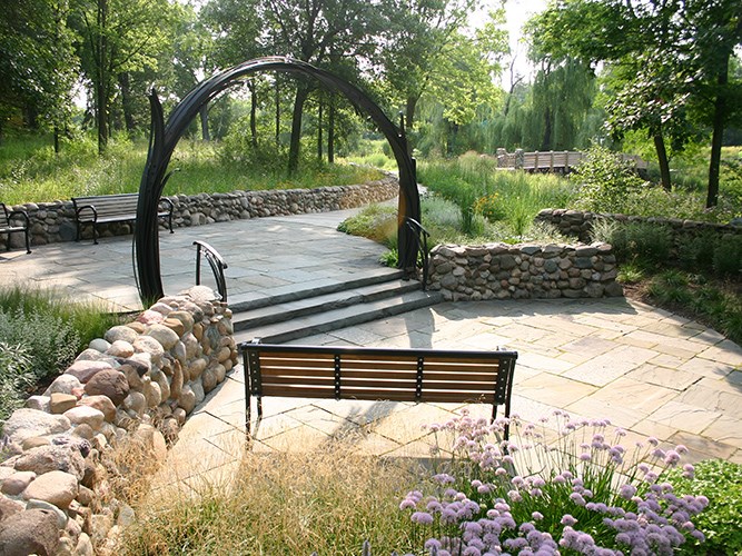 A curved stone pathway leads up shallow steps to a circular metal arch set among benches, wildflowers, and trees in a landscaped garden.
