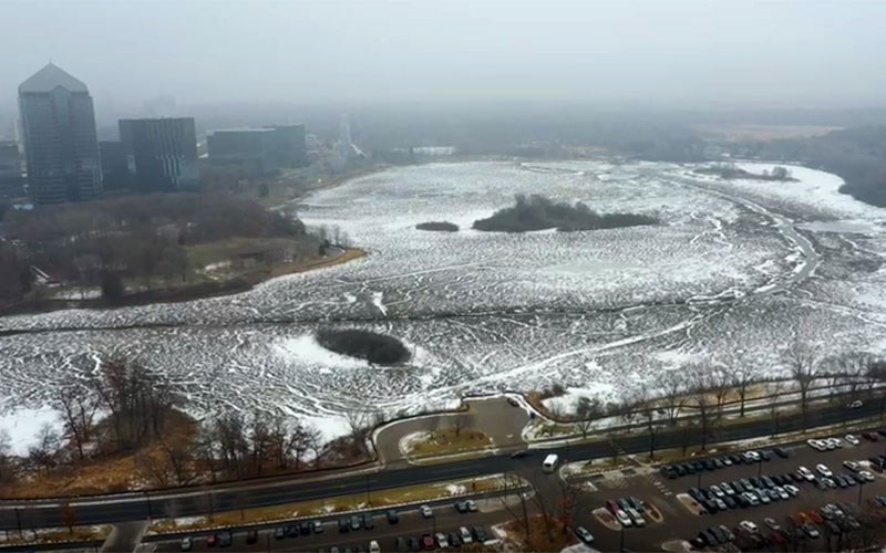 A foggy aerial view shows a snow-covered wetland surrounded by leafless trees, nearby roads, and several large buildings in the distance.