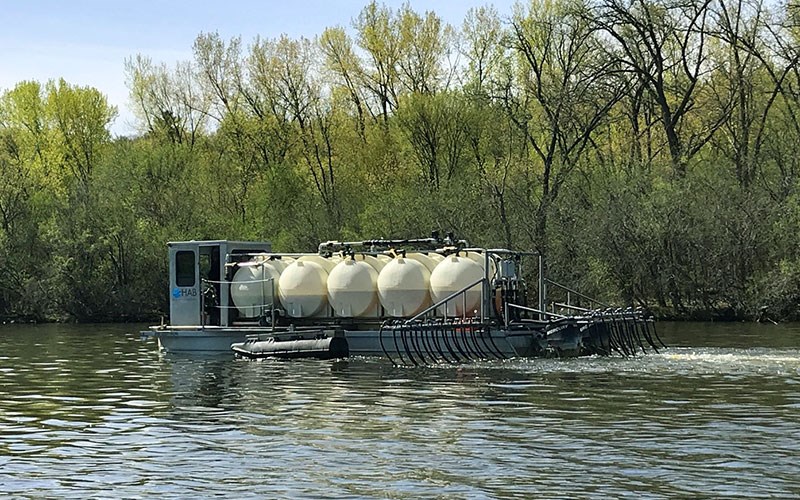 A metal work boat carrying several large white tanks moves across a calm body of water with dense green trees in the background.