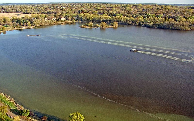 A small boat creates a curving wake as it moves across a large lake surrounded by dense spring foliage and a few small islands.