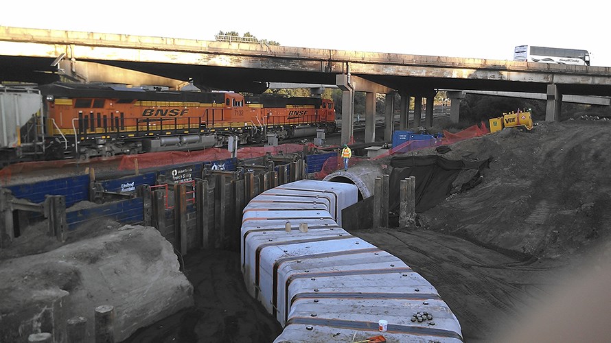 An orange BNSF train passes behind a construction site where large concrete tunnel segments curve through an excavated trench beneath an elevated highway.