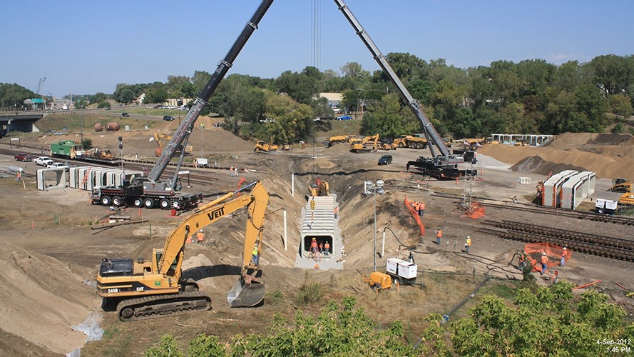 Construction crews, cranes, and excavators work around a large trench and rail lines on a wide, busy job site surrounded by trees.