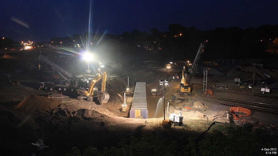 Bright work lights illuminate construction vehicles and crews working around a large trench and rail lines on a nighttime job site.