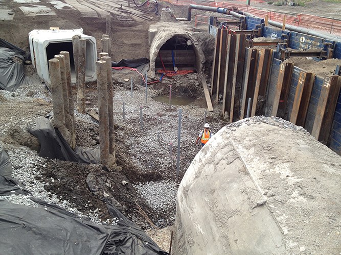 An excavated construction site shows exposed soil walls, concrete tunnel sections, vertical support piles, and a worker standing near gravel‑covered ground.