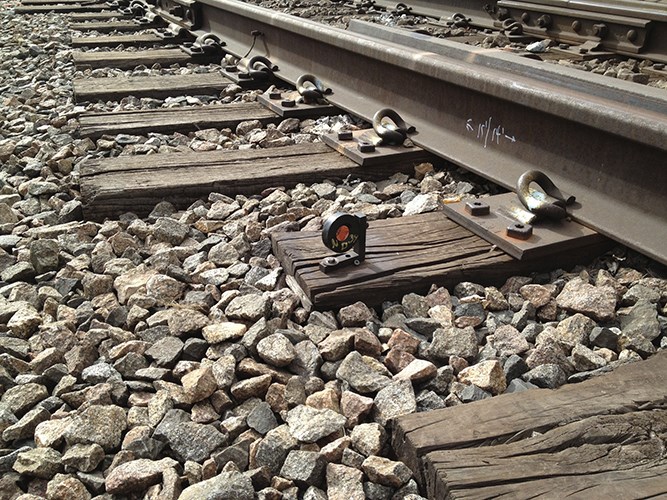 A small measuring device rests on a wooden railroad tie among gravel beside steel train tracks.