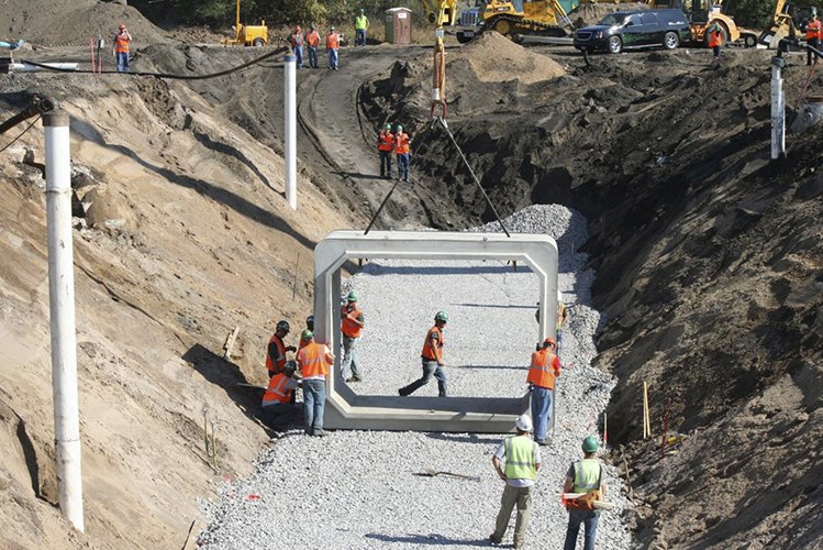 Construction workers guide a large square concrete tunnel segment being lowered into a gravel-lined trench between steep dirt walls.