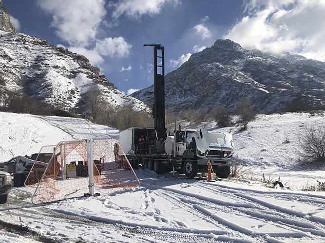 A drilling truck and equipment sit on snowy ground at the base of rugged, snow‑dusted mountains under a partly cloudy sky.