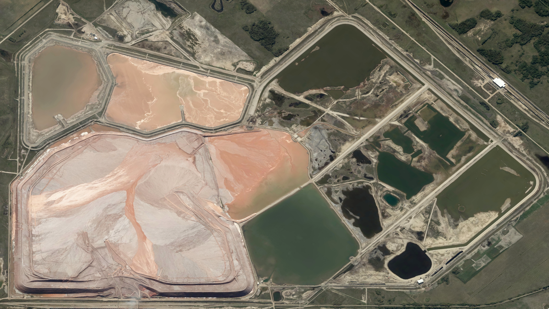 Aerial view of a potash mine site with multiple containment ponds of varying colors—green, brown, and reddish—surrounded by dirt roads and sparse vegetation.